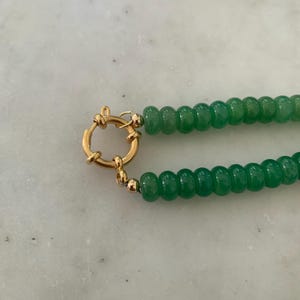 May include: A necklace featuring two strands of small, round, vibrant green beads. The strands are connected by a circular gold-toned spring ring clasp, which is attached to the beads with small gold-toned metal beads at each end.