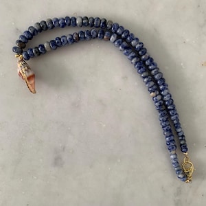 May include: A necklace featuring two strands of small, round, mottled blue and white beads. A single, small, spiral-shaped seashell charm with a gold-toned metal cap hangs from one end, and a gold-toned metal lobster claw clasp secures the other end.