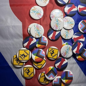 May include: A collection of circular pin-back buttons featuring various designs, including white buttons with the text GOOD PUP, yellow diamond-shaped signs with the text GOOD BOY, and buttons displaying colorful striped patterns and red bone icons.