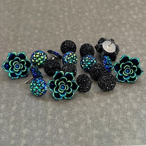 May include: An assortment of earrings in various styles. The collection includes black, sparkly stud earrings, iridescent blue and green stud earrings, and flower-shaped earrings with a blue and green metallic finish. The earrings are displayed on a gray surface.