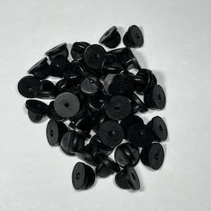 May include: A pile of black rubber earring backs. The backs have a disc shape with a small hole in the center and a tapered post. The earring backs are scattered on a white surface.