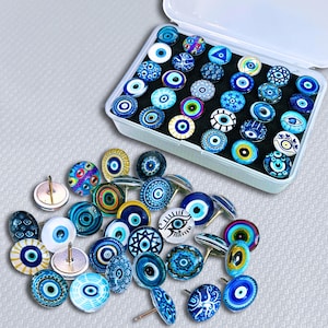 May include: A collection of blue and white Evil Eye themed push pins and cufflinks. The pins and cufflinks feature various designs, including concentric circles, eye motifs, and geometric patterns. Some are stored in a white plastic container.