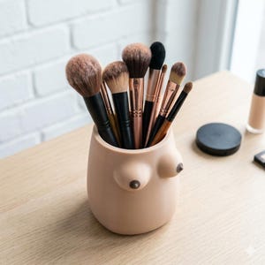 May include: A beige, breast-shaped makeup brush holder filled with various makeup brushes. The holder has two prominent breast-like shapes with dark brown nipples. The brushes have black and rose gold handles and various brush heads.