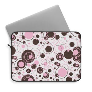May include: A laptop sleeve featuring a repeating pattern of solid and outlined circles in shades of dark brown and light pink on a white background. The sleeve is rectangular with a black zipper edge, partially containing a silver laptop.