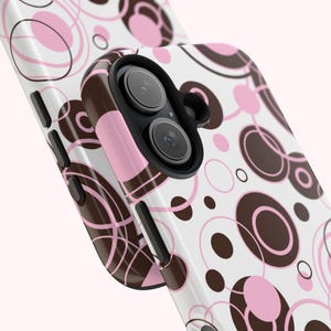 May include: A white smartphone case featuring a repeating pattern of overlapping circles in shades of dark brown and light pink. The case has a black camera lens cutout and black side buttons, showcasing a modern geometric design on the back surface.