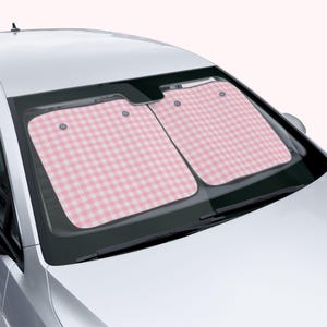 May include: A two-piece car windshield sun shade featuring a light pink and white gingham checkered pattern. The rectangular panels are positioned inside the front window of a silver vehicle, covering the glass to block sunlight from the interior.