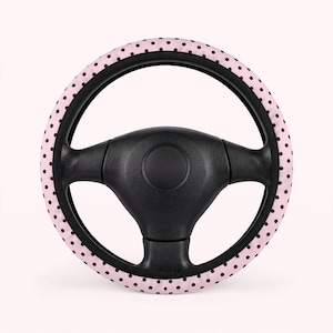 May include: A black steering wheel with a pink cover featuring a pattern of small black polka dots. The steering wheel cover is a circular shape and fits over the wheel. The background is a light pink color.
