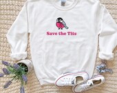 Photo 5 of 20: A white crewneck sweatshirt featuring a bird graphic with a pink ribbon and the text Save the Tits is displayed flat with white sneakers, lavender flowers, and a scrunchie on a knitted background.
