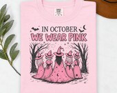 Photo 2 of 17: A folded light pink t-shirt with a witch graphic and In October We Wear Pink text, displayed on a white surface with foliage and a brown hat.