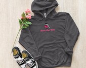 Photo 10 of 20: A charcoal gray hoodie featuring a bird graphic with a pink ribbon and the text Save the Tits is laid flat on a wooden background with black sneakers and pink roses.