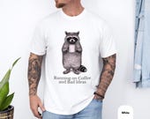 Photo 4 of 8: Charcoal unisex raccoon shirt laid on rustic wood, featuring a hand drawn raccoon clutching a steaming coffee mug and subtle "Running on Coffee and Bad Ideas" text, funny raccoon tee with sarcastic coffee humor for introverts, cozy soft cotton look, novelty gift for him
