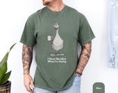 Photo 5 of 8: Folded charcoal Comfort Colors tee on rustic wood featuring a soft vintage-style goose illustration holding a coffee mug, subtly distressed print and cozy worn-in feel, playful animal humor and introvert sarcasm, ideal funny goose shirt or funny graphic tee as a gift for him, dad, husband, or mama t-shirt