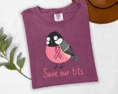 Photo 4 of 17: A folded maroon t-shirt with a printed cartoon bird design and pink text reading Save our tits.