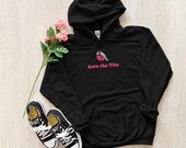 Photo 8 of 20: A black hoodie featuring a graphic of a bird with a pink ribbon and the text Save the Tits is laid out on a light wood surface alongside black and white sneakers and pink roses.