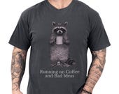 Photo 1 of 8: Black unisex t shirt on a tattooed male model showing a detailed raccoon illustration holding a coffee mug and a cheeky slogan about being fueled by coffee and questionable decisions, a raccoon shirt and funny raccoon tee with coffee humor and introvert-friendly novelty vibe, cozy soft cotton gift for him