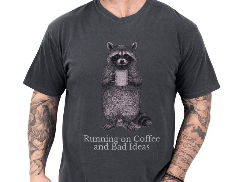 Black unisex t shirt on a tattooed male model showing a detailed raccoon illustration holding a coffee mug and a cheeky slogan about being fueled by coffee and questionable decisions, a raccoon shirt and funny raccoon tee with coffee humor and introvert-friendly novelty vibe, cozy soft cotton gift for him