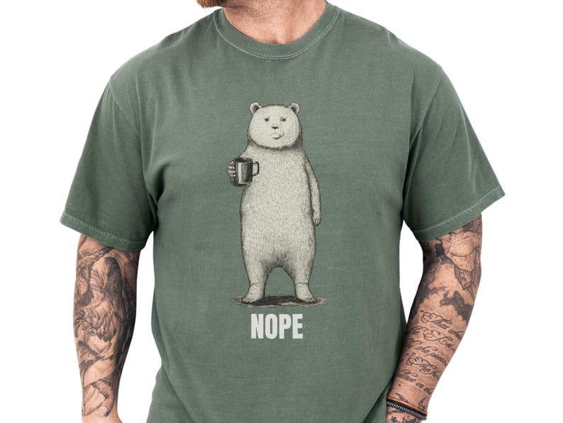 White unisex funny bear shirt showing a sketched bear holding a coffee mug with bold NOPE caption, a minimalist sarcastic t shirt perfect for introvert humor and coffee humor shirt lovers, styled on a tattooed model for cozy everyday novelty graphic tee gift for him