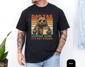 Photo 3 of 8: Man wearing a soft blue jean garment-dyed unisex dad t-shirt showing a retro sunset graphic of a sunglasses-wearing bear holding fries and a soda with bold DAD TAX text — cozy, casual dad joke tee and funny dad shirt ideal as a fathers day tee or dad birthday gift