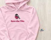 Photo 1 of 20: A light pink hoodie with a printed graphic of a bird with a pink ribbon and the text Save the Tits lies flat on a wooden surface.
