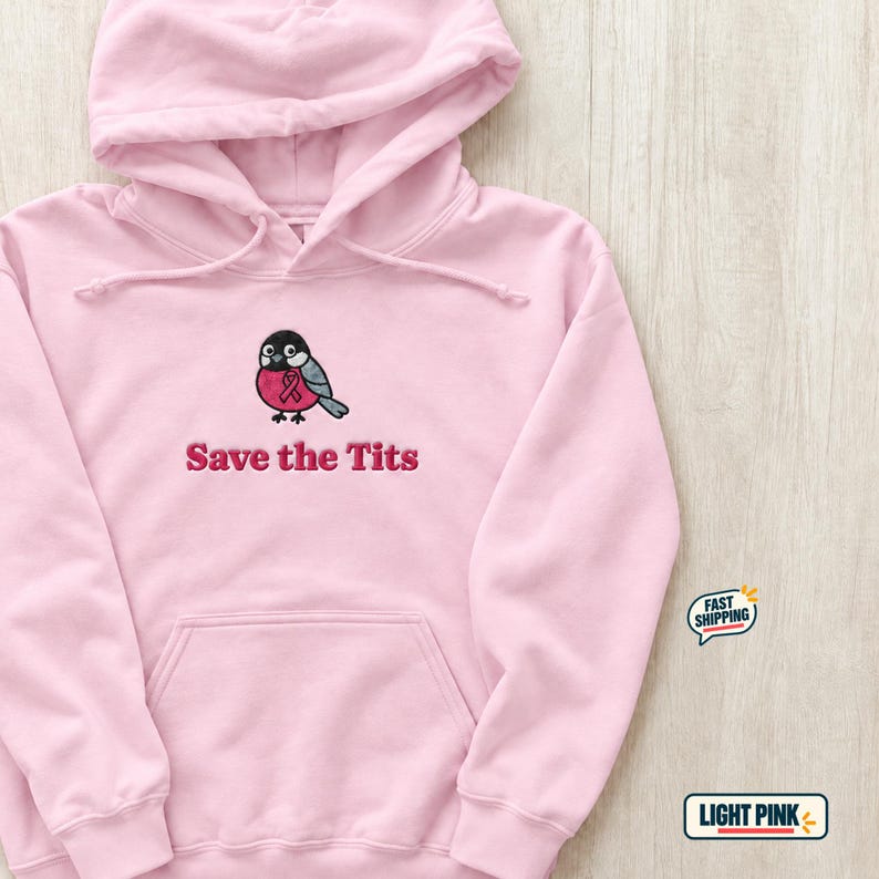 A light pink hoodie with a printed graphic of a bird with a pink ribbon and the text Save the Tits lies flat on a wooden surface.