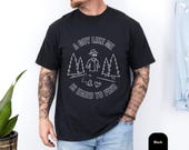 Photo 3 of 8: Outdoorsy man wearing a muted olive Comfort Colors cotton sarcastic tee featuring a playful "a guy like me" invisible man camping graphic, leaning on a mountain bike on a leafy trail, cozy worn-in feel ideal funny dad joke tee gift for cyclists and hikers
