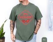 Photo 5 of 8: Casual moss green tee with bold red "0% VEGAN" sign and steak icons, soft vintage wash and relaxed fit shown with jeans and tattoos for a backyard grilling mood — ideal funny BBQ shirt or grilling tee, backyard bbq tee, meat lover gift or grillmaster shirt for dad