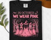 Photo 4 of 17: A folded black t-shirt printed with the In October We Wear Pink witch design, placed on a white background with a green sprig and brown hat.