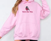 Photo 3 of 20: A person wears an oversized light pink hoodie featuring a bird graphic with a pink ribbon and the text Save the Tits.