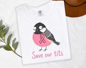 Photo 9 of 17: A folded white t-shirt displaying a cartoon bird graphic with a pink ribbon and text.