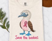Photo 8 of 17: A folded cream-colored t-shirt featuring a blue-footed booby bird graphic with a pink ribbon and the text Save the boobies.