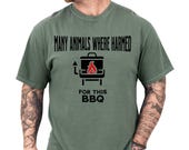 Photo 1 of 8: Olive green short-sleeve tee on a tattooed model featuring a bold black grill icon with flame and the cheeky typo line "Many animals where harmed for this BBQ" — playful many animals shirt and typo shirt that doubles as a meme tshirt, dark humor tshirt, funny bbq shirt, grilling shirt, barbecue tee, ideal sarcastic shirt and bbq gift idea for a meat lover at casual cookouts with a rustic, laid-back vibe