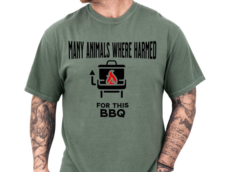 Olive green short-sleeve tee on a tattooed model featuring a bold black grill icon with flame and the cheeky typo line "Many animals where harmed for this BBQ" — playful many animals shirt and typo shirt that doubles as a meme tshirt, dark humor tshirt, funny bbq shirt, grilling shirt, barbecue tee, ideal sarcastic shirt and bbq gift idea for a meat lover at casual cookouts with a rustic, laid-back vibe