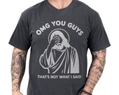 Photo 1 of 8: Charcoal vintage-wash unisex tee showing a distressed Jesus facepalm illustration with bold "OMG YOU GUYS" and the caption "That's not what I said", a funny Jesus shirt and meme graphic tee with cozy worn-in feel and an edgy, sarcastic Christian humor vibe perfect as a gift for friends