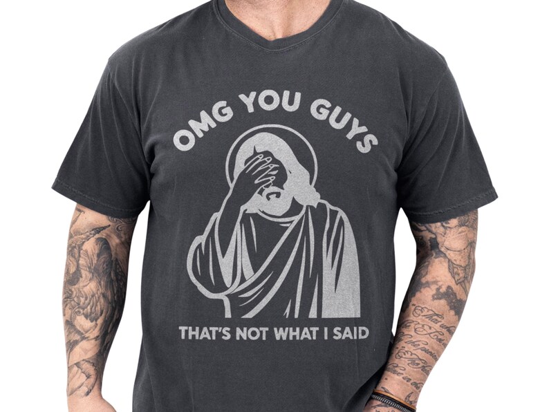 Charcoal vintage-wash unisex tee showing a distressed Jesus facepalm illustration with bold "OMG YOU GUYS" and the caption "That's not what I said", a funny Jesus shirt and meme graphic tee with cozy worn-in feel and an edgy, sarcastic Christian humor vibe perfect as a gift for friends