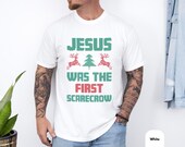 Photo 4 of 7: Funny christian tee with a retro holiday graphic reading Jesus was the first scarecrow, styled like a cozy ugly sweater design with festive trees and reindeer, a witty jesus scarecrow tee for Christmas gifting or faith-filled laughs