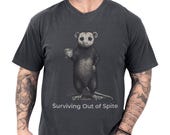 Photo 1 of 8: Funny opossum tee on a charcoal comfort colors shirt with a dark, cozy look, featuring a quirky possum holding a drink and the phrase Surviving Out of Spite, a sarcastic gift for him or anyone who loves introvert humor and animal humor shirt style