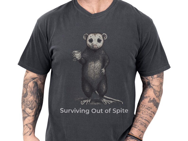 Funny opossum tee on a charcoal comfort colors shirt with a dark, cozy look, featuring a quirky possum holding a drink and the phrase Surviving Out of Spite, a sarcastic gift for him or anyone who loves introvert humor and animal humor shirt style