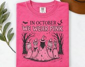Photo 3 of 17: A folded dark pink t-shirt featuring the In October We Wear Pink witch graphic, resting on a white surface next to leaves and a hat.