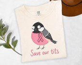 Photo 8 of 17: A folded off-white t-shirt featuring a cartoon bird illustration and pink text.