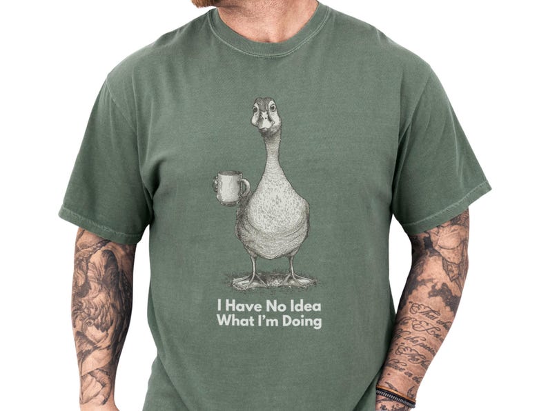 Casual man models a moss green unisex funny goose shirt featuring a sketched goose holding a coffee mug and the caption I Have No Idea What I'm Doing, a soft, worn-in funny graphic tee with meme t shirt vibes, sarcastic novelty goose t shirt and animal humor shirt, great gift for him or gift for dad, cozy introvert humor apparel