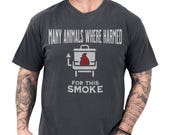 Photo 1 of 8: Charcoal vintage-washed tee on a tattooed model showing a cheeky smoker graphic with red flame and distressed slogan "Many animals where harmed for this smoke", a bbq humor shirt and dark humor tee perfect as a funny grill shirt or meat smoker gift for a grillmaster dad