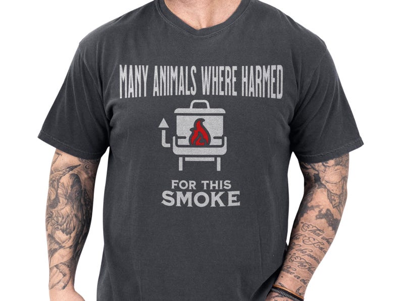 Charcoal vintage-washed tee on a tattooed model showing a cheeky smoker graphic with red flame and distressed slogan "Many animals where harmed for this smoke", a bbq humor shirt and dark humor tee perfect as a funny grill shirt or meat smoker gift for a grillmaster dad