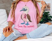 Photo 2 of 17: A woman sitting on a white bed wearing a pink t-shirt with a blue-footed booby bird graphic and pink ribbon.