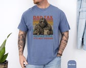 Photo 6 of 8: Casual cyclist on a sun-dappled forest trail wearing a Dad Tax t shirt featuring a retro Sasquatch silhouette and sunset stripes, a funny dad shirt and sasquatch shirt with vintage vibe, perfect camping dad shirt and outdoor dad gift for birthdays or weekends