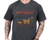 Photo 1 of 10: Black unisex funny shirt on a male model showing pixel art ox and covered wagon by trees and mountains, bold orange PLEASE BE PATIENT headline with smaller I was born in the 1900s text, a sarcastic graphic t and funny retro tee with retro gaming vibe, ideal novelty joke tshirt gift for Gen X and millennial gamers