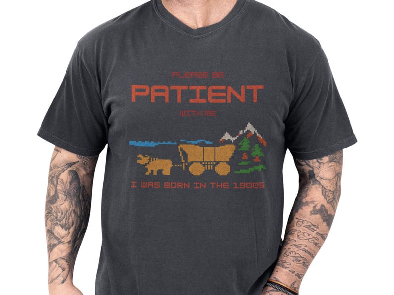 Black unisex funny shirt on a male model showing pixel art ox and covered wagon by trees and mountains, bold orange PLEASE BE PATIENT headline with smaller I was born in the 1900s text, a sarcastic graphic t and funny retro tee with retro gaming vibe, ideal novelty joke tshirt gift for Gen X and millennial gamers