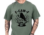 Photo 1 of 8: Unisex crow shirt in muted green with a bold black raven perched over a skull and bare branches, featuring the playful F-CAW-F design for fans of dark humor tshirt style and gothic crow tee looks, great as a sarcastic tee or gift for men