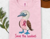 Photo 5 of 17: A folded light pink t-shirt featuring a blue-footed booby bird graphic with a pink ribbon and text that reads Save the boobies.