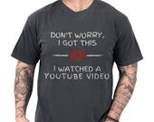 Photo 1 of 8: Black soft cotton funny dad tee featuring a distressed retro play icon and bold hand-lettered slogan "Don't worry I got this — I watched a YouTube video", a playful YouTube repair joke shown on a tattooed handyman, cozy mens humor apparel ideal as a fathers day present or birthday gag gift for the garage project guy