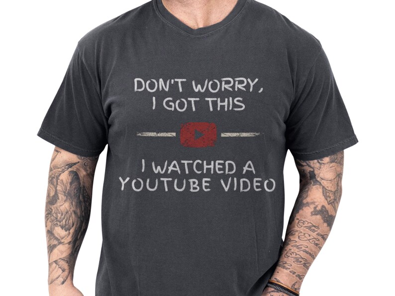 Black soft cotton funny dad tee featuring a distressed retro play icon and bold hand-lettered slogan "Don't worry I got this — I watched a YouTube video", a playful YouTube repair joke shown on a tattooed handyman, cozy mens humor apparel ideal as a fathers day present or birthday gag gift for the garage project guy
